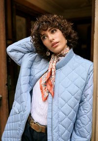 Ženska kodrastih las, oblečena v svetlo modro prešito jakno, z vzorčasto rutko, zlatimi uhani in pasom, pozira z roko za glavo ob vratah.