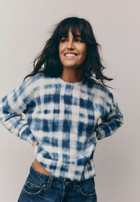 Pull en tricot avec un motif tie-dye dans des nuances de bleu et blanc, présentant un col rond et des poignets côtelés, porté avec un jean en denim.