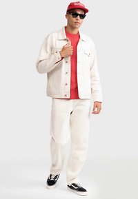 Bijela traper jakna preko crvenog džempera s pletenim uzorkom, bijele udobne hlače i crne tenisice. Dodaci uključuju crvenu kapu i sunčane naočale.