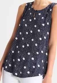 Top sin mangas de color navy con lunares blancos, que presenta un detalle en forma de ojo de cerradura en el cuello y un corte relajado con un dobladillo ligeramente acampanado.