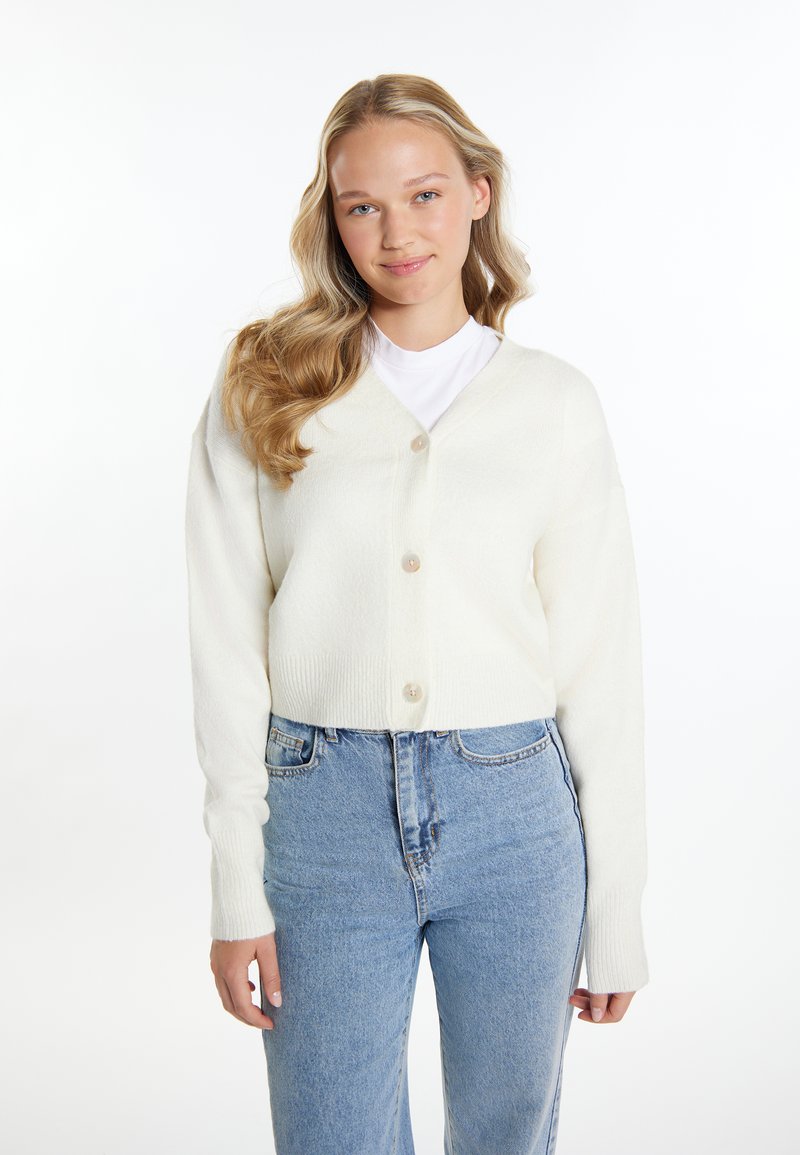 Weißer, gestrickter, verkürzter Cardigan mit V-Ausschnitt und fünf Knöpfen. Lange Ärmel, weicher Stoff und gerippte Bündchen, kombiniert mit hoch taillierten Jeans.