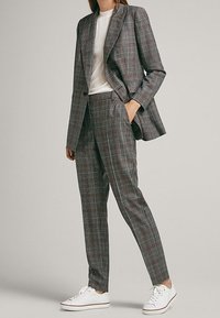 Femme portant un costume à carreaux gris avec un t-shirt blanc et des baskets blanches, se tenant avec une main dans la poche devant un fond uni.