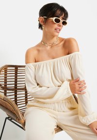 Cremfarbener Off-Shoulder-Top aus strukturiertem Stoff mit schmal zulaufenden Ärmeln; kombiniert mit runden Sonnenbrillen und geschichtetem Schmuck, sitzend auf einem geflochtenen Stuhl.