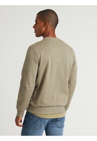 Sudadera de algodón verde con puños y bajo de canalé. Diseño de cuello redondo. La vista trasera muestra detalles de costura. Combinada con jeans de mezclilla azules.