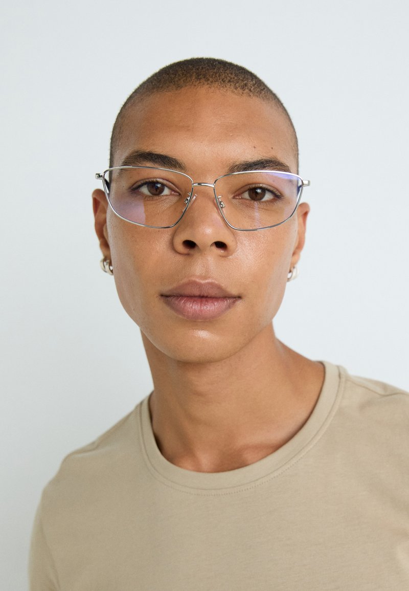 Des lunettes en métal avec des verres rectangulaires, dotées d'une finition argentée et de branches transparentes. Portées par une personne portant un t-shirt beige.