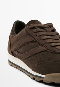 Brown suede sneaker with a textured upper, featuring curved stitching details, a padded collar, and a white midsole with a contrasting outsole.