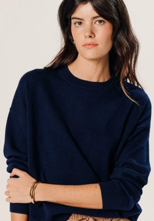 Frau mit dunklen Haaren, die einen marineblauen Pullover mit langen Ärmeln und goldene Armbänder trägt und posiert, während sie einen Arm über ihren Oberkörper verschränkt.