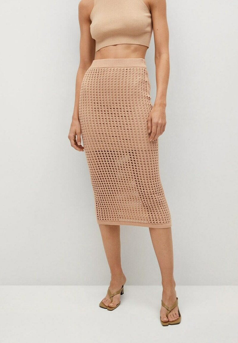 Femme portant un haut court beige et une jupe midi en maille filet beige, avec des talons ouverts, debout contre un mur uni.