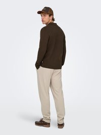 Brown long-sleeve sweater, beige pants, and brown shoes; a brown cap with a mesh back. The figure stands with hands in pockets, viewed from behind.