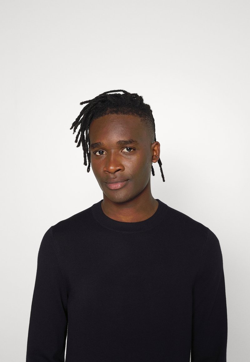 Jonge zwarte man met korte dreadlocks in een zwarte ronde hals trui, staande tegen een effen lichtgrijze achtergrond.