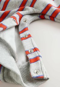 Sivý pletený odev s červenými a modrými horizontálnymi pruhmi, s kovovými patentkami pozdĺž výstrihu. Viditeľná mäkká vnútorná podšívka.