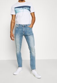 Bílé tričko s modrým grafickým nápisem "JACK&JONES", doplněné o světle modré úzké džíny a bílé tenisky, stojící před jednobarevným pozadím.