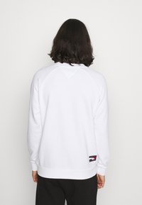 Person med axellångt mörkt hår som bär en vit sweatshirt med en liten rektangulär logotyp nära nedre delen av ryggen, svarta byxor, stående.