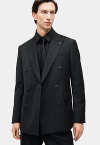 Νέος άνδρας που φοράει εφαρμοστό μαύρο διπλό σακάκι με διακριτικές λεπτές ρίγες, μαύρο πουκάμισο και μαύρη γραβάτα, κοιτάζει προς τα πλάγια.