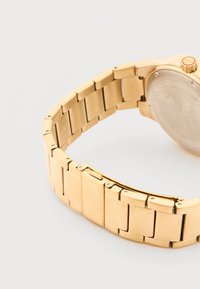 Χρυστό μεταλλικό λουράκι ρολογιού με συνδεδεμένα τμήματα και αγκράφα, προσαρτημένο σε κάσα ρολογιού με κορώνα σημειωμένη "HUGO".