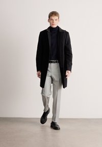 Casaco de lã preto por cima de uma camisola de gola alta azul-marinho, combinado com calças cinza claro e sapatos pretos. Texturas suaves, design minimalista.