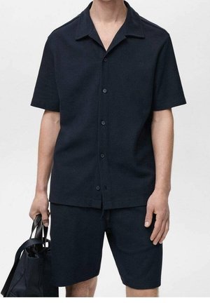 Mężczyzna w czarnej koszuli na krótki rękaw zapinanej na guziki oraz czarnych spodenkach, trzymający czarną torbę typu tote w lewej ręce.