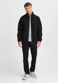 Chaqueta de forro polar negra con un cuello alto, superficie texturizada y cremallera frontal. Llevada sobre una camiseta gris clara con pantalones negros y zapatos deportivos.
