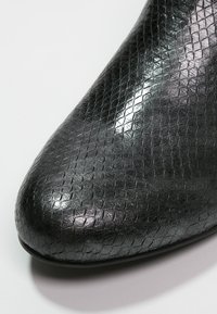 Svart lädersko med en texturerad, präglad yta som liknar fjäll. Har en spetsig tå och en glansig, reflekterande yta.