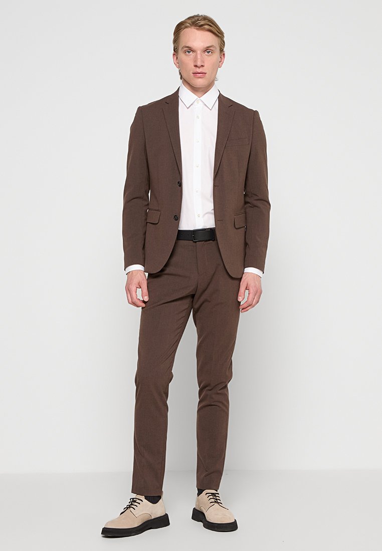 Traje marrón con una camisa blanca, diseño ajustado, textura suave y dos bolsillos frontales. Complementado con zapatos beige claro con cordones.