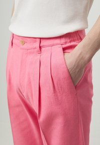 Różowe bawełniane spodnie z przodu z plisami, zapięciem na guzik i bocznymi kieszeniami. Tkanina wydaje się gładka z lekkim połyskiem.