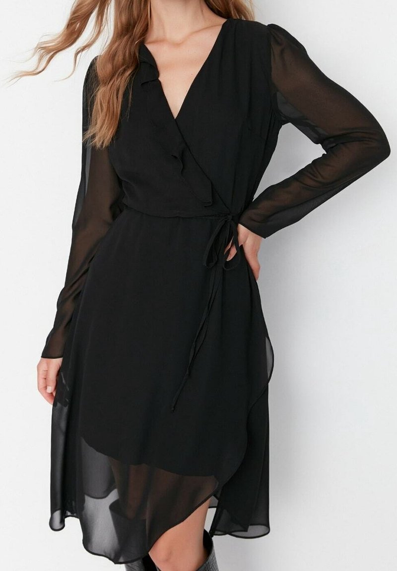 Robe portefeuille noire avec manches longues transparentes, col à volants et taille nouée. L'ourlet est asymétrique, créant une silhouette fluide.