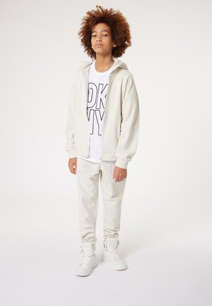 Lichtbeige hoodie met trekkoord en voorrits, gecombineerd met bijpassende joggers. Het witte T-shirt eronder heeft vetgedrukte zwarte tekst. Witte sneakers.