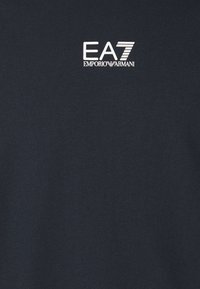 Tamnobla bomba majica iz bombaža z belim logotipom "EA7" in "EMPORIO ARMANI" v velikih črkah, postavljena na zgornji levi strani. Gladka tekstura.