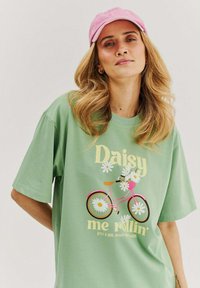 Woman wearing a pink cap and green t-shirt with a pink bicycle and daisy flowers graphic that says "Daisy me rollin'".