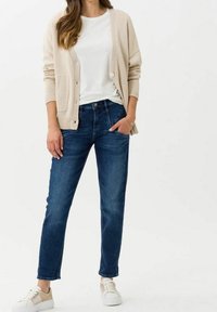 Beige pletený cardigan, bílý tričko s kulatým výstřihem, tmavě modré džíny rovného střihu a bílé tenisky s růžovými tkaničkami.
