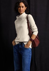 Frau mit kurzen lockigen Haaren, die einen cremefarbenen Strickpullover, blaue Jeans und ein rotes Lederarmband trägt und eine rote Lederumhängetasche hält.