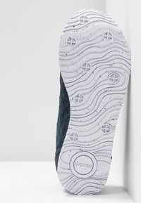 Semelle de chaussure avec un motif ondulé blanc, des éléments de traction circulaires, et le nom de la marque « WODEN » sur une tige en tissu texturé foncé.