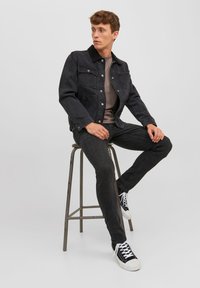 Jachetă din denim negru cu închidere cu nasturi și două buzunare la piept, asociată cu un tricou gri și blugi negri. Tenisi negri și albi completează look-ul.