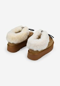 Un par de zapatillas de plataforma color beige con un grueso forro de felpa blanca y lazos negros, sobre un fondo blanco.