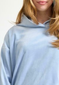 Jasnoniebieska bluza z kapturem, wykonana z miękkiego materiału; charakteryzuje się luźnym krojem i regulowanym sznurkiem w kapturze. Brak widocznych wzorów lub akcentów.