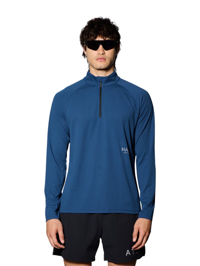 Man draagt een blauw atletisch shirt met lange mouwen en rits, zwarte shorts en zwarte om de ogen geslagen zonnebril, staand tegen een effen witte achtergrond.
