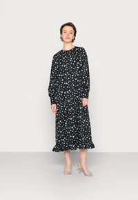 Vestido preto de mangas compridas com bolinhas azul claro, com bainha em babados e um corte solto. Modelo a usar sapatos de salto azuis claros correspondentes.