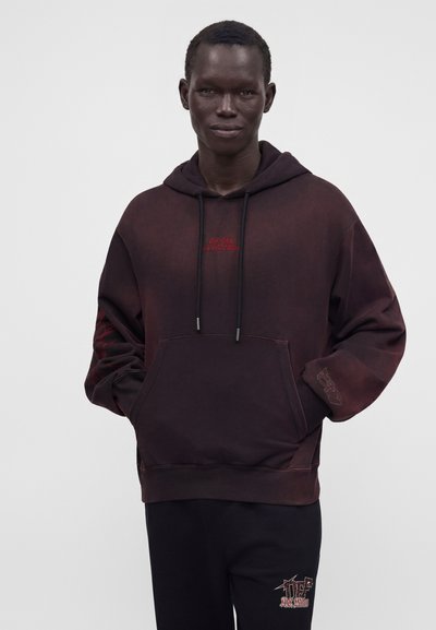 Homme portant un sweat à capuche sombre surdimensionné avec un texte rouge sur la poitrine et la manche, et un pantalon noir avec un logo rouge et blanc.