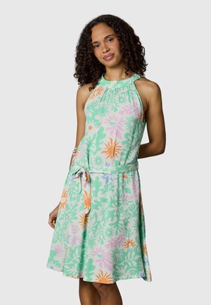 Kvinne med krøllete hår iført en ermeløs, knelang blomsterkjole med grønne, oransje og rosa mønstre, stående mot en ensfarget bakgrunn.