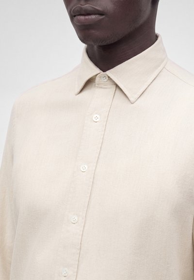 Gros plan d'une personne portant une chemise beige boutonnée avec un col pointu, se concentrant sur le haut de la poitrine et la zone du cou.