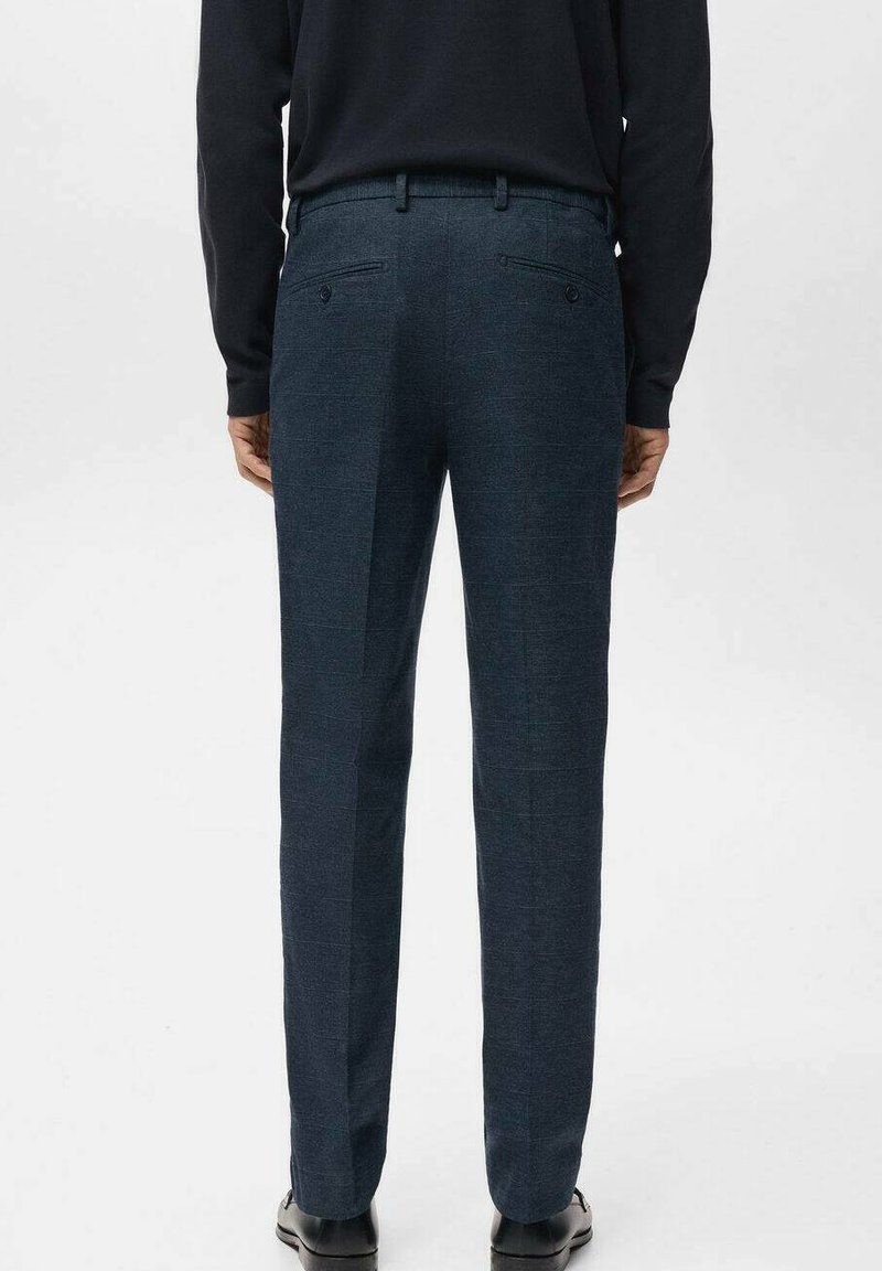 Homme portant un pantalon habillé bleu foncé, une chemise à manches longues noire et des chaussures noires, debout les mains le long du corps, de dos.