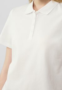 Witte poloshirt met een klassieke kraag, korte mouwen en knoopsluiting. Materiaal lijkt zacht met een gladde textuur.