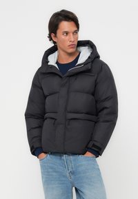 Doudoune noire avec capuche, dotée d'une doublure grise, fermeture zippée à l'avant, poches à rabat et poignets élastiques. Texture lisse avec un design matelassé.