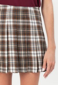 Brown and white plaid skirt with pleats, featuring a textured fabric and a fitted waist. Hand resting on the edge with a solid color top visible.