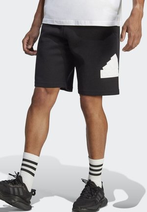 Pantalones cortos negros de corte holgado, con un logo blanco de Adidas en el lado izquierdo. Combinados con zapatillas negras y calcetines a rayas blancas.