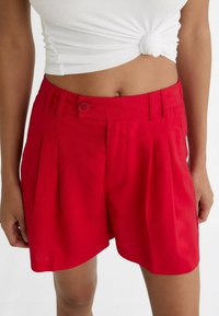 Shorts plissés rouges avec une fermeture par bouton, design ajusté et texture douce. Le tissu est léger et vibrant.