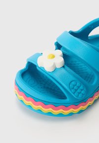 Sininen kumisaapas, jossa on kolme remmiä ja valkoinen kukkaaksentti. Ominaisuuksina monivärinen aaltoileva pohja, jossa on vaaleanpunaista, keltaista ja sinistä tekstuuria.