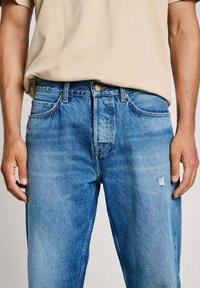 Sinise denimist valmistatud teksad, millel on mugav lõige, paremal jalal kulunud plaastriga, messingist detailid ja klassikaline viie tasku disain.