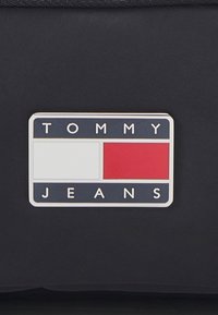 Logo Tommy Jeans, prostokątne z granatowym tłem, białymi i czerwonymi kwadratami oraz białym tekstem. Plastikowy materiał o gładkiej fakturze.