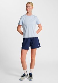 T-shirt de sport bleu clair à manches courtes associé à un short marine. Le t-shirt a un col rond et un logo subtil. Le modèle porte des baskets grises et des chaussettes blanches.